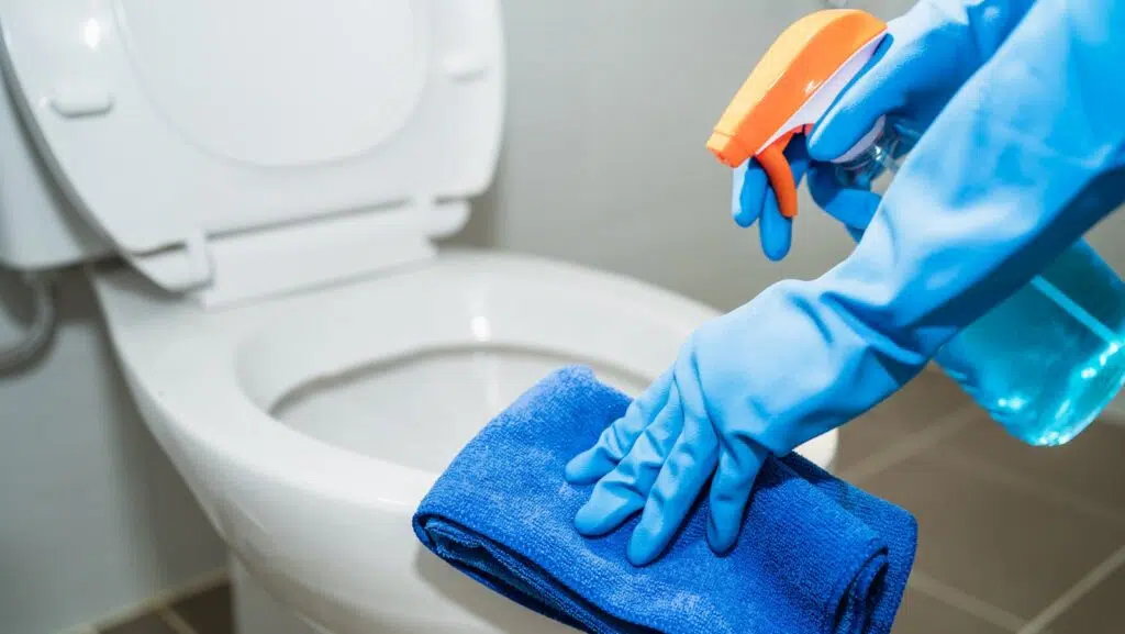 Professional cleaner in blue rubber gloves disinfecting a commercial toilet with spray bottle and microfiber cloth